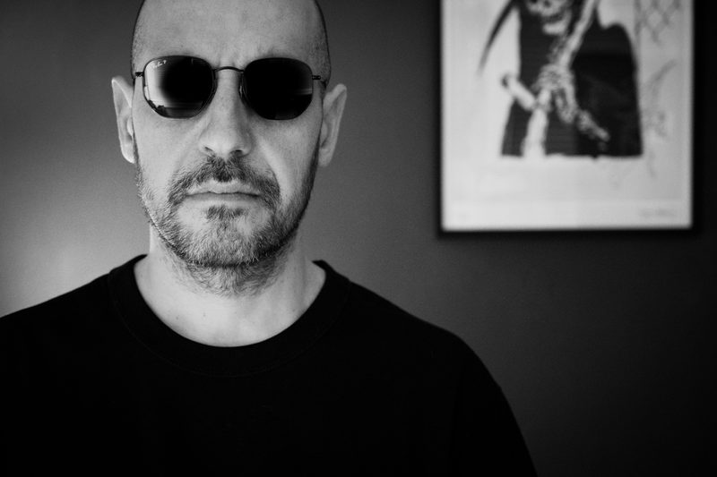 Black and white portrait of Danyboy wearing sunglasses, a framed print on the wall behind him