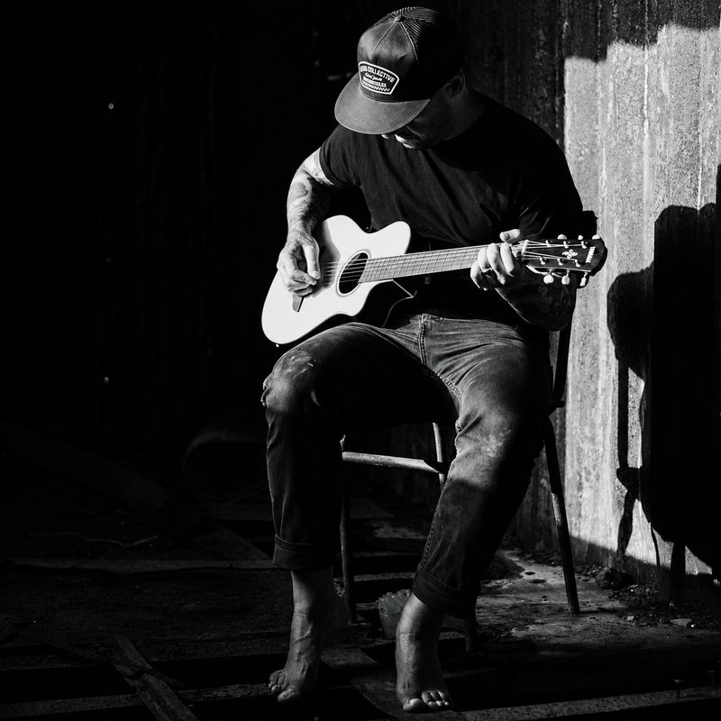 Black and white photograph of Danyboy seated on a stool playing an acoustic guitar in a dark room