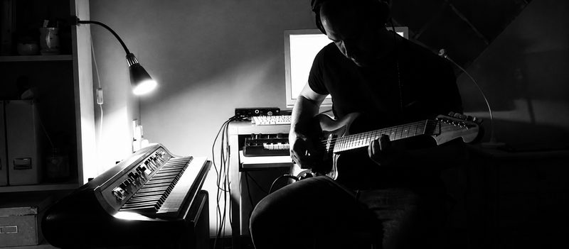Black and white photograph of Danyboy playing electric guitar in a home studio beside a lamp-lit organ and synth rack
